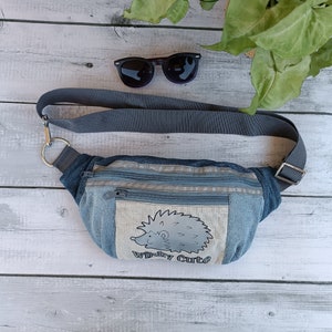 Puede incluir: Una riñonera de mezclilla gris con un bolsillo con cremallera y un parche blanco con un erizo de dibujos animados con el texto "Wildly Cute".