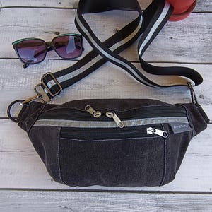May include: A black denim fanny pack with a silver reflective strip and two zipped compartments. The bag has a black adjustable strap with silver accents. The bag has a label that reads "Anne Sather".