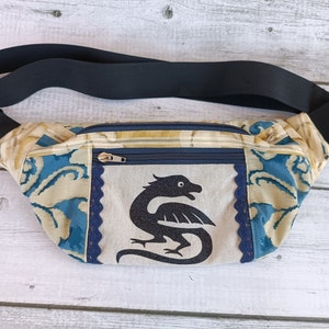 May include: A blue and cream fabric fanny pack with a black dragon design. The pack has a zipper closure and a black adjustable strap.