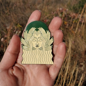 Può includere: Spilla in smalto color oro raffigurante il volto di una donna con capelli fluenti e ali piumate. La spilla ha un alone verde scuro e un viso e capelli color crema. La spilla è tenuta in mano su uno sfondo sfocato di erba secca.