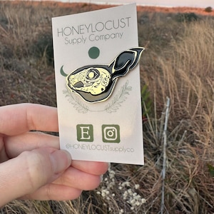 Pu&ograve; includere: Una spilla in smalto nero e oro con un design a teschio di coniglio. La spilla &egrave; attaccata a una carta con il testo "HONEYLOCUST Supply Company" e i social media. Lo sfondo mostra un campo di erba alta e un tramonto.