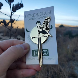 May include: A gold-colored metal pin featuring a black bird perched on a sword. The pin is on a white card with the text "HONEYLOCUST Supply Company" and social media handles. The bird has detailed wing feathers.