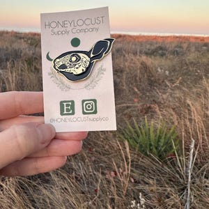 Pu&ograve; includere: Una spilla in smalto nero e oro a forma di teschio di coniglio, esposta su una carta con il testo "HONEYLOCUST Supply Company". La spilla presenta dettagli intricati ed &egrave; tenuta contro uno sfondo di erba alta e un tramonto.