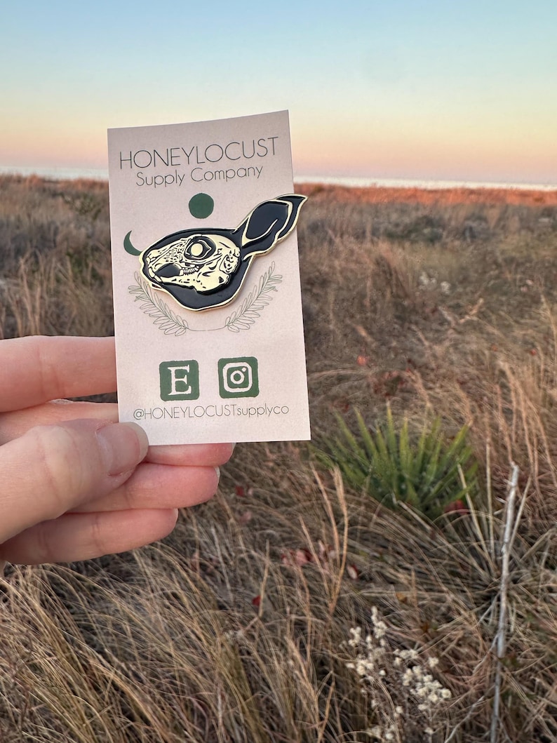 Pu&ograve; includere: Una spilla in smalto nero e oro a forma di teschio di coniglio, esposta su una carta con il testo "HONEYLOCUST Supply Company". La spilla presenta dettagli intricati ed &egrave; tenuta contro uno sfondo di erba alta e un tramonto.