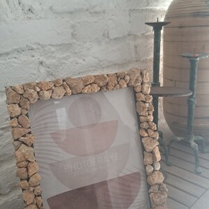 Puede incluir: Un marco de fotos rústico hecho de pequeñas piedras, con una impresión abstracta marrón y rosa en el interior. El marco mide 20x25cm y se encuentra sobre una superficie de madera.