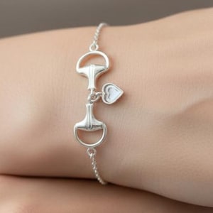 May include: A silver bracelet featuring a horse bit design with a small heart charm. The bracelet is delicate and has a chain link style. The horse bit is a classic equestrian symbol.