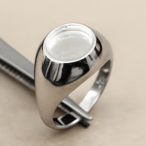Puede incluir: Un anillo de sello plateado pulido con una cara ovalada lisa, sostenido por unas pinzas. El anillo tiene un diseño clásico, ideal para la personalización. El anillo está hecho de un metal brillante y reflectante.