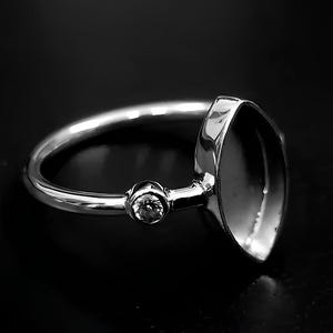 Könnte beinhalten: Ein silberner Ring mit einer Lünettenfassung und einem kleinen, runden Akzentstein. Der Ring hat ein dünnes, poliertes Band. Der Ring wird auf einer dunklen Oberfläche präsentiert.
