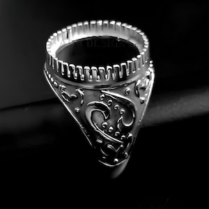 Puede incluir: Un anillo plateado con un diseño ornamentado. El anillo presenta un patrón detallado en la banda y un engaste circular en la parte superior. El anillo se encuentra sobre un fondo oscuro, resaltando sus intrincados detalles.
