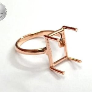 Puede incluir: Anillo de oro rosa con engaste rectangular para una piedra preciosa. El anillo es de plata de ley 925, como indica el sello circular del fondo. El anillo está sobre un fondo blanco.