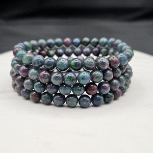 May include: A multi-strand bracelet composed of round, polished beads in shades of deep blue, forest green, and dark magenta. The beads are strung together in a circular formation, creating a layered appearance with a smooth, reflective surface texture.