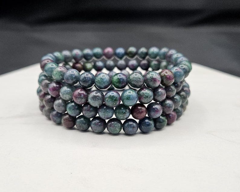 May include: A multi-strand bracelet composed of round, polished beads in shades of deep blue, forest green, and dark magenta. The beads are strung together in a circular formation, creating a layered appearance with a smooth, reflective surface texture.