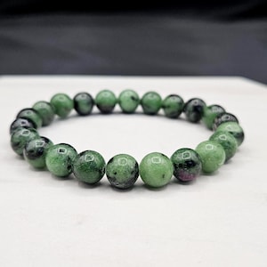 May include: A stretch bracelet composed of round, polished beads in shades of mottled green and black, with occasional small inclusions of deep magenta or dark red, arranged in a continuous circular loop on a smooth, light-colored surface.