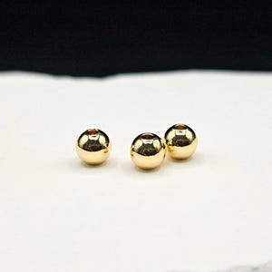 May include: Three small, spherical, metallic gold-colored beads with a smooth, reflective surface. Each bead features a central hole for threading, showcasing a polished finish that catches the light, creating bright highlights on their rounded forms.