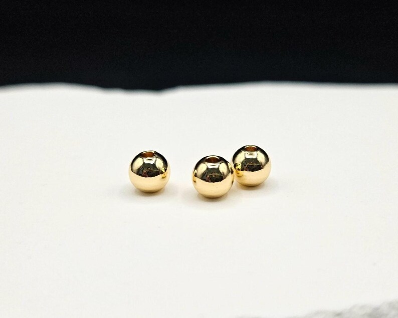 May include: Three small, spherical, metallic gold-colored beads with a smooth, reflective surface. Each bead features a central hole for threading, showcasing a polished finish that catches the light, creating bright highlights on their rounded forms.