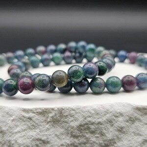 May include: A beaded bracelet featuring smooth, round stones in a variegated palette of deep forest green, royal blue, and dark magenta. The polished spheres are strung together in a continuous loop, showcasing natural color variations and a glossy finish.