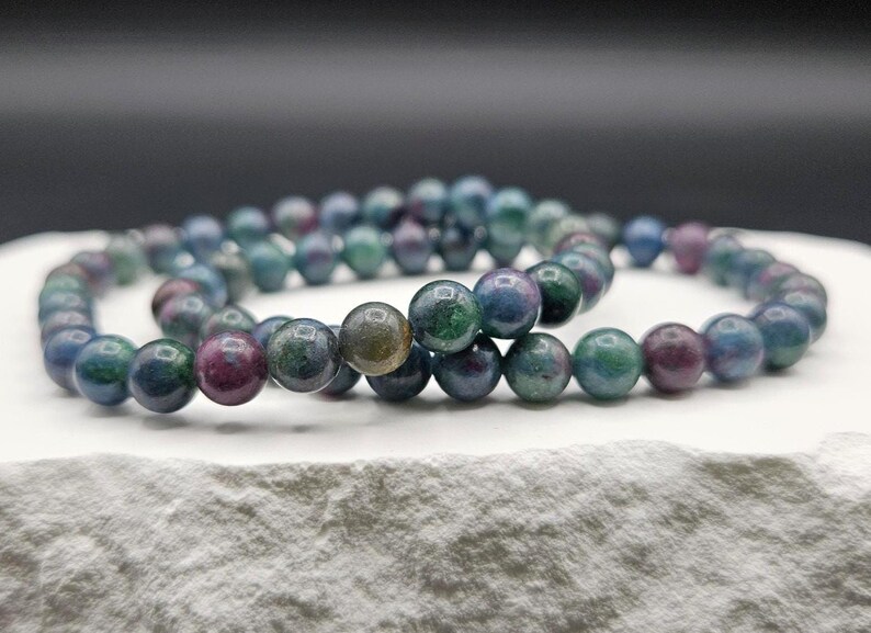 May include: A beaded bracelet featuring smooth, round stones in a variegated palette of deep forest green, royal blue, and dark magenta. The polished spheres are strung together in a continuous loop, showcasing natural color variations and a glossy finish.