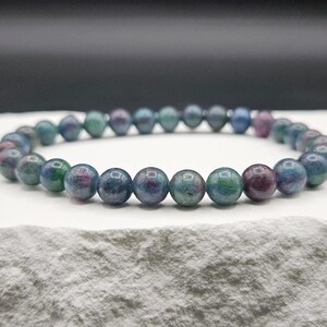 May include: A beaded bracelet featuring smooth, round stones in variegated shades of deep blue, teal, and magenta. The beads are strung together in a continuous circular loop, showcasing a polished, reflective surface with natural color variations.