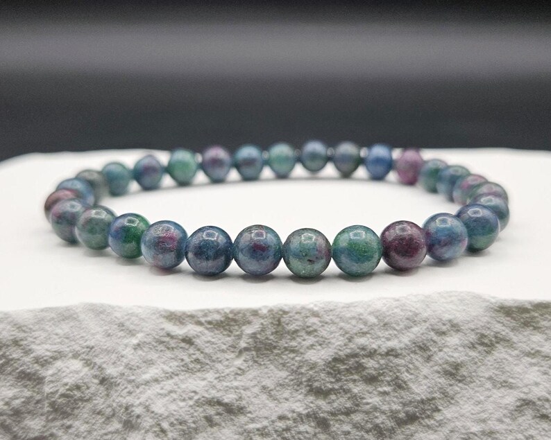 May include: A beaded bracelet featuring smooth, round stones in variegated shades of deep blue, teal, and magenta. The beads are strung together in a continuous circular loop, showcasing a polished, reflective surface with natural color variations.