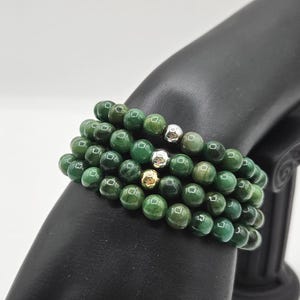 Peut inclure: Un bracelet multi-rangs composé de perles rondes et polies de couleur verte avec des motifs marbrés. Le bracelet comprend trois perles d'accent métalliques distinctes : une argentée, une dorée et une argentée texturée, alignées en rangée.