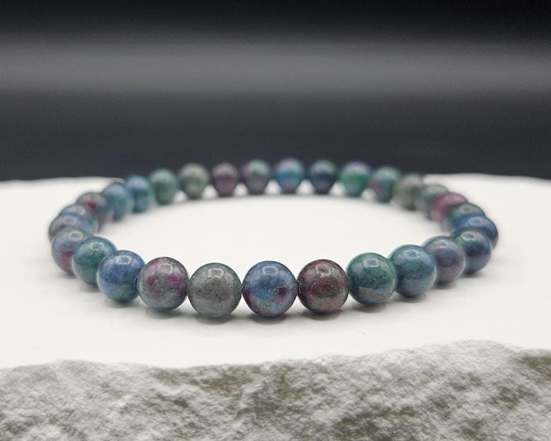 May include: A stretch bracelet composed of smooth, round beads in varying shades of deep teal, blue, and muted burgundy. The beads are polished to a glossy finish and are strung together in a continuous circular loop, creating a simple jewelry piece.