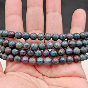May include: A multi-strand beaded bracelet featuring round, polished gemstone beads in variegated shades of deep blue, forest green, and magenta. The beads are strung together in a continuous circular pattern, showcasing a natural, mottled texture.
