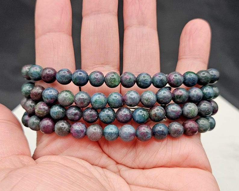 May include: A multi-strand beaded bracelet featuring round, polished gemstone beads in variegated shades of deep blue, forest green, and magenta. The beads are strung together in a continuous circular pattern, showcasing a natural, mottled texture.