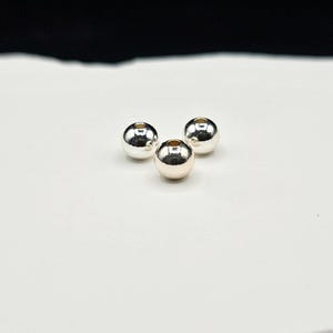 May include: Three small, spherical, metallic silver beads with a smooth, reflective surface. Each bead features a centered circular hole, indicating they are designed for stringing. The beads are arranged in a triangular formation.
