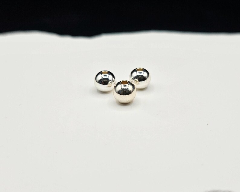 May include: Three small, spherical, metallic silver beads with a smooth, reflective surface. Each bead features a centered circular hole, indicating they are designed for stringing. The beads are arranged in a triangular formation.