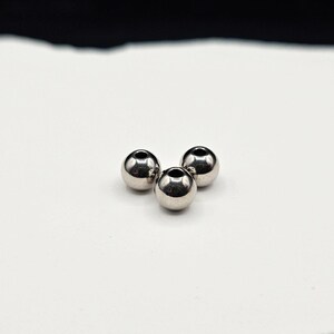 May include: Three small, spherical, polished silver-toned metal beads arranged in a triangular cluster. Each bead features a smooth, reflective surface and a centered circular hole, creating a uniform appearance for these metallic jewelry components.