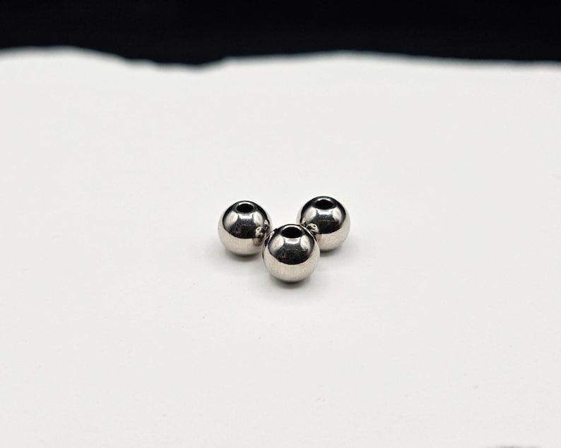 May include: Three small, spherical, polished silver-toned metal beads arranged in a triangular cluster. Each bead features a smooth, reflective surface and a centered circular hole, creating a uniform appearance for these metallic jewelry components.