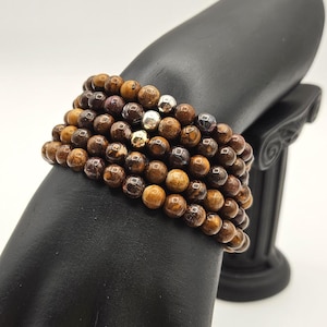 May include: A multi-strand bracelet composed of numerous round, polished brown and tan beads with a mottled, earthy texture. The design features several small, reflective metallic accent beads in silver and gold tones interspersed throughout the rows.