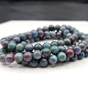 May include: A beaded bracelet composed of multiple strands of polished, spherical gemstones. The beads feature a mottled color palette of deep forest green, muted teal, and dark magenta, with a smooth, reflective surface texture throughout.