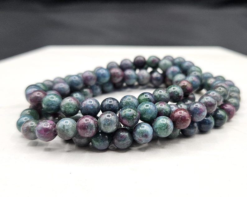 May include: A beaded bracelet composed of multiple strands of polished, spherical gemstones. The beads feature a mottled color palette of deep forest green, muted teal, and dark magenta, with a smooth, reflective surface texture throughout.