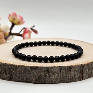 May include: A circular bracelet composed of small, smooth, polished black spherical beads strung together. The beads are uniform in size and color, creating a continuous loop. The bracelet is presented as a single strand of dark, reflective stones.