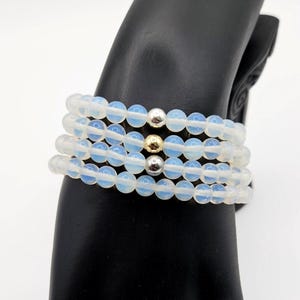 May include: A set of four stretch bracelets featuring translucent, milky white opal-like beads. Each bracelet is accented with a single metallic bead, alternating between polished silver-tone and gold-tone spheres, arranged in a stacked formation.