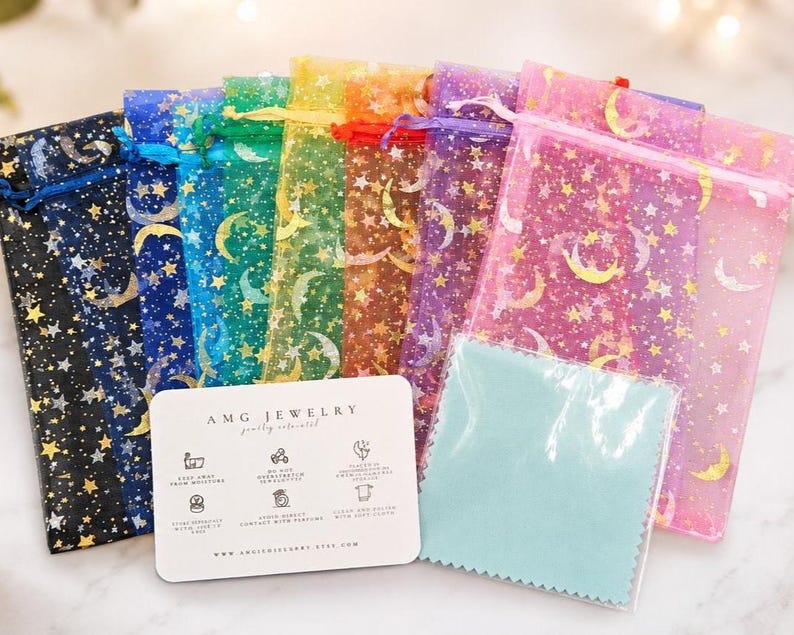 May include: A collection of sheer organza drawstring pouches in black, blue, green, yellow, orange, purple, and pink, each featuring a gold foil moon and star pattern. A white rectangular jewelry care card and a light blue polishing cloth are included.