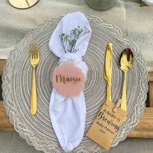Könnte beinhalten: Eine weiße Serviette mit einer rosafarbenen Acryl-Namensschilder, auf der "Maisie" steht. Die Serviette liegt auf einem goldenen Platzdeckchen mit einer goldenen Gabel und einem Messer. Eine braune Papiertüte mit dem Text "A Baby is Brewing Baby Palmer 2021" liegt auf dem Tisch.