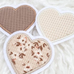 Könnte beinhalten: Drei herzförmige Aufnäher mit weißen Rändern. Einer ist einfarbig braun, ein weiterer cremefarben mit einem strukturierten Muster, und der dritte hat ein Blumenmuster in Braun und Creme.