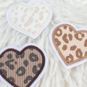 Könnte beinhalten: Drei herzförmige Aufnäher mit Leopardenmuster. Jeder Aufnäher hat einen weißen Rand und ein anderes Farbschema: Creme, Braun und Beige. Die Aufnäher sind auf einer flauschigen weißen Oberfläche angeordnet.