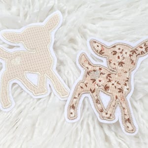 Könnte beinhalten: Zwei hirschförmige Aufnäher. Einer ist beige mit strukturiertem Strick-Aussehen, der andere hat ein Blumenmuster in Braun- und Beigetönen. Beide Aufnäher haben einen weißen Rand und sind vor einem flauschigen weißen Hintergrund platziert.