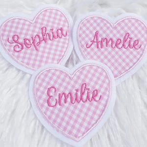 Könnte beinhalten: Drei herzförmige Aufnäher mit weißen Rändern. Jeder Aufnäher hat ein rosa-weißes Vichy-Muster und einen Namen, der in rosa Schrift gestickt ist: Sophia, Amelie und Emilie. Die Aufnäher sind auf einer weißen, flauschigen Oberfläche angeordnet.