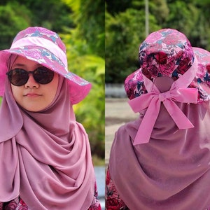 Puede incluir: Un sombrero de cubo floral rosa con un lazo de cinta rosa en la parte posterior. El sombrero está hecho de una tela suave y ligera y tiene un ala ancha.
