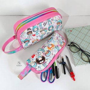 Może przedstawiać: Dwa różowe piórniki z kolorowym nadrukiem kreskówek pielęgniarek. Większy piórnik ma zamek błyskawiczny i uchwyt. Mniejszy piórnik ma zamek błyskawiczny i uchwyt. Oba piórniki mają nadrukowany tekst "Our Heroes" i "Handmade". Mniejszy piórnik jest wypełniony ołówkami, długopisami, kompasem i nożyczkami.