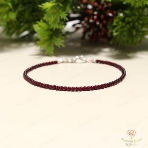 Peut inclure: Un bracelet délicat orné de petites perles rondes, rouge foncé. Le bracelet est doté d'un fermoir argenté et est présenté sur une surface claire, avec un feuillage vert en arrière-plan. Les perles semblent être des pierres précieuses.