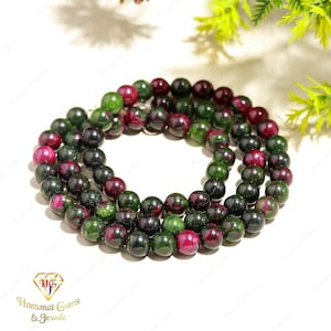 May include: A circular strand of polished, spherical beads featuring a mottled pattern of deep forest green and vibrant magenta-red. The beads are arranged in a continuous loop, showcasing a smooth, reflective surface with natural color variations.