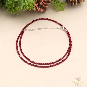 May include: A delicate necklace featuring small, faceted, deep red beads. The necklace is double-stranded and has a silver-colored chain and clasp. The beads are evenly spaced, creating a refined and elegant look. The background is a soft, neutral color.