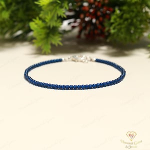 May include: A fine bracelet composed of small, round, dark blue beads. The bracelet is secured with a silver-coloured clasp. The jewellery is presented on a light surface, with green foliage visible in the background.