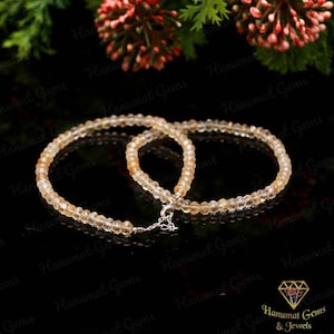 Op de afbeelding: Een ketting bestaande uit kleine, ronde, licht amberkleurige kralen. De ketting is vastgemaakt met een zilveren sluiting. De achtergrond is zwart, met bloemaccenten aan de bovenkant. De ketting is waarschijnlijk een sieraad.