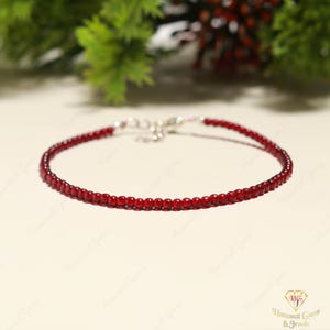 May include: A fine red beaded bracelet with a silver clasp. The bracelet is composed of small, round, deep red beads. The background features greenery and a soft, neutral surface.